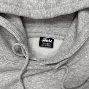 May include: Close-up of a light gray hoodie with a drawstring and hood. The Stussy logo is visible on a tag inside the hood. The hoodie is a size XL.