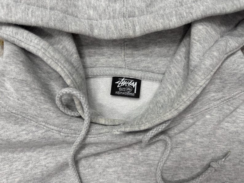 May include: Close-up of a light gray hoodie with a drawstring and hood. The Stussy logo is visible on a tag inside the hood. The hoodie is a size XL.