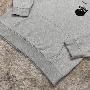 May include: A light gray hoodie with a kangaroo pocket and a drawstring hood. The hoodie features a black and white graphic of an eight ball on the upper left chest. The cuffs and hem are ribbed.
