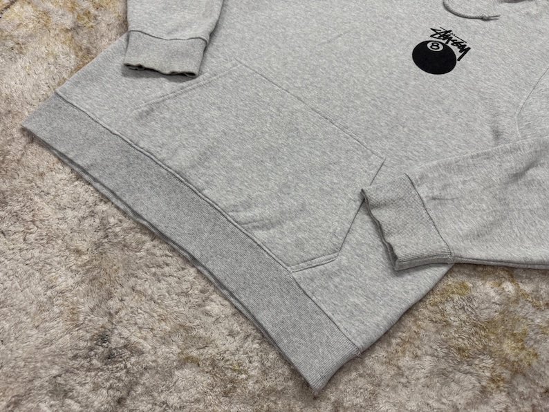 May include: A light gray hoodie with a kangaroo pocket and a drawstring hood. The hoodie features a black and white graphic of an eight ball on the upper left chest. The cuffs and hem are ribbed.