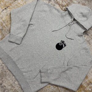 May include: A heather gray hoodie with a drawstring hood and a kangaroo pocket. The hoodie features a black graphic of an eight ball with the word "Stussy" in a stylized font.