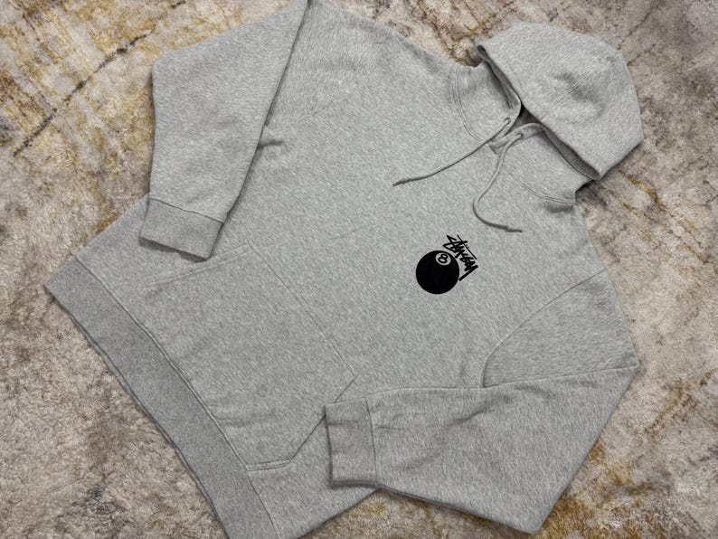 May include: A heather gray hoodie with a drawstring hood and a kangaroo pocket. The hoodie features a black graphic of an eight ball with the word "Stussy" in a stylized font.