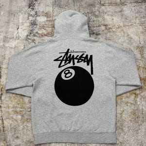 May include: A gray hoodie featuring the St&uuml;ssy logo and an 8-ball graphic in black on the back. The hoodie has a hood and a kangaroo pocket. The fabric appears to be a soft, comfortable material.