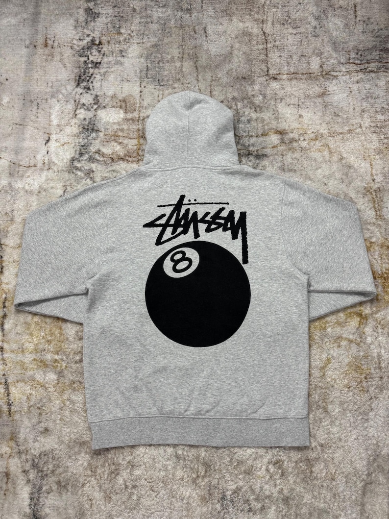 May include: A gray hoodie featuring the St&uuml;ssy logo and an 8-ball graphic in black on the back. The hoodie has a hood and a kangaroo pocket. The fabric appears to be a soft, comfortable material.
