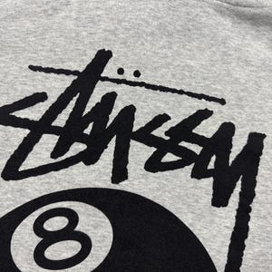 May include: Close-up of a gray sweatshirt featuring a large black "Stussy" logo above a black 8-ball graphic. The design is centered on the fabric, showcasing the brand's iconic style. The fabric has a soft, textured appearance.