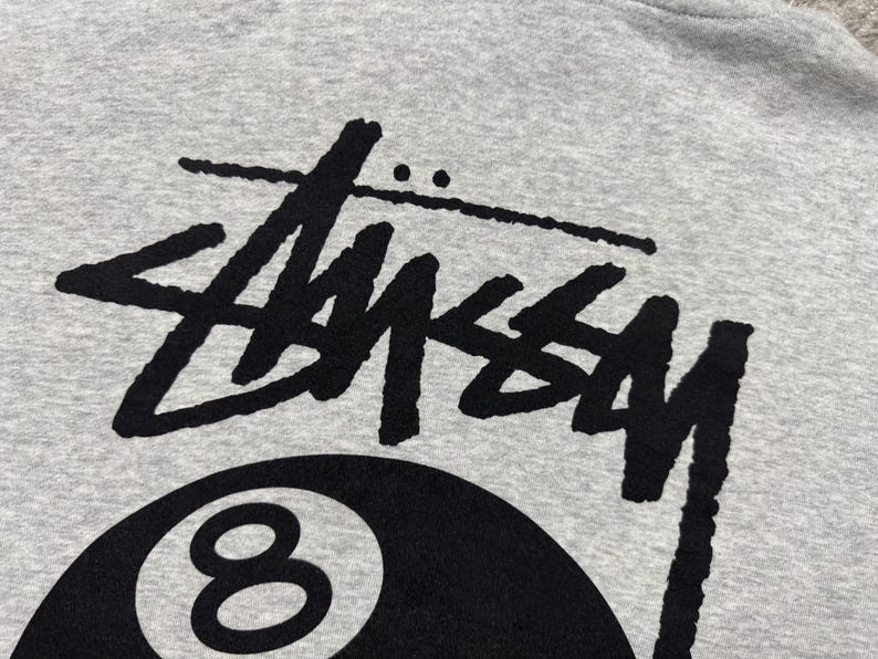 May include: Close-up of a gray sweatshirt featuring a large black "Stussy" logo above a black 8-ball graphic. The design is centered on the fabric, showcasing the brand's iconic style. The fabric has a soft, textured appearance.