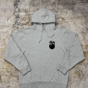 May include: A heather gray hoodie with a drawstring hood and a kangaroo pocket. The hoodie features a black graphic of an eight ball and the Stussy logo on the front.