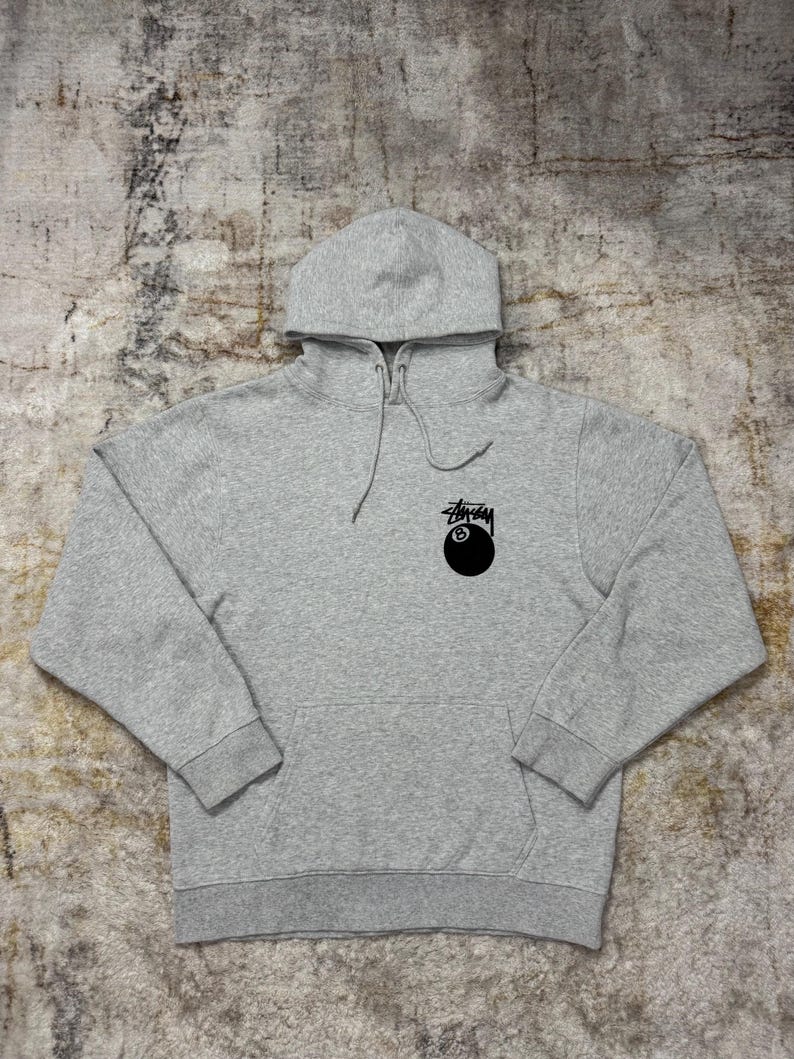 May include: A heather gray hoodie with a drawstring hood and a kangaroo pocket. The hoodie features a black graphic of an eight ball and the Stussy logo on the front.