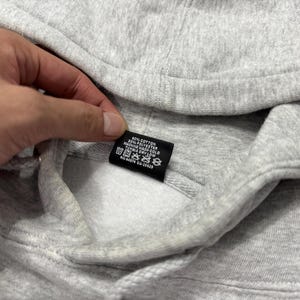 May include: Close-up of a gray hooded sweatshirt. The sweatshirt has a drawstring and a tag with care instructions. The tag is black with white text. The fabric is a soft, textured material.