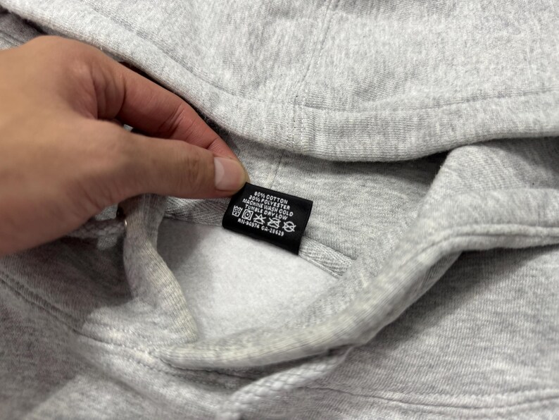 May include: Close-up of a gray hooded sweatshirt. The sweatshirt has a drawstring and a tag with care instructions. The tag is black with white text. The fabric is a soft, textured material.