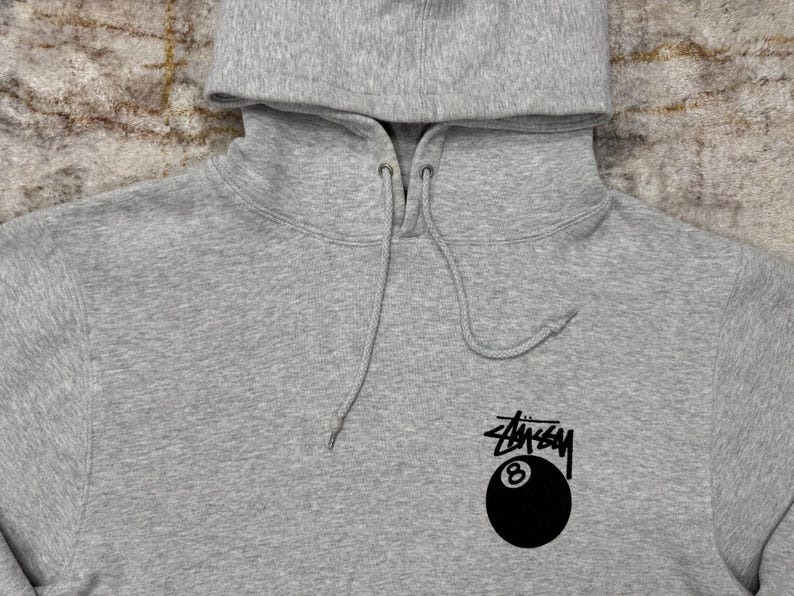 May include: A heather gray hoodie with a drawstring hood. The hoodie features a black and white logo with the number 8 inside a black eight ball.