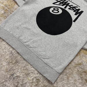 May include: A gray sweatshirt featuring the word "STUSSY" in black block letters above a large black eight ball graphic. The sweatshirt has a ribbed hem and cuffs.