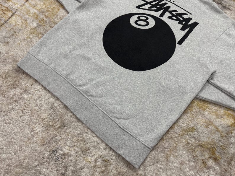 May include: A gray sweatshirt featuring the word "STUSSY" in black block letters above a large black eight ball graphic. The sweatshirt has a ribbed hem and cuffs.