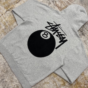May include: A heather gray hoodie featuring a large black 8-ball graphic and the Stussy logo. The hoodie has a hood and long sleeves. The design is printed on the back of the hoodie.
