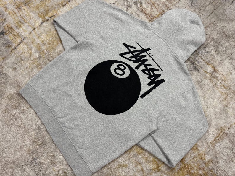 May include: A heather gray hoodie featuring a large black 8-ball graphic and the Stussy logo. The hoodie has a hood and long sleeves. The design is printed on the back of the hoodie.