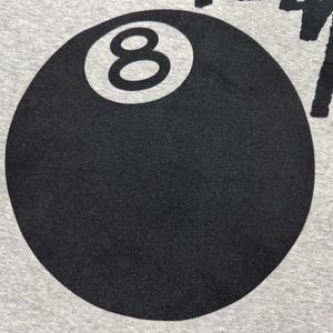 May include: Close-up of a gray fabric with a large black 8-ball graphic. The number 8 is in white within the black circle. The word "Stussy" is printed in black on the top right.