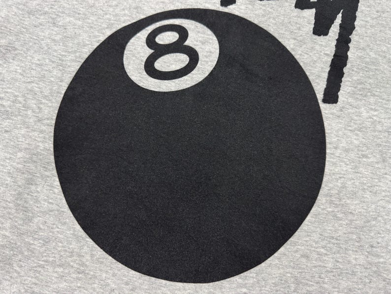 May include: Close-up of a gray fabric with a large black 8-ball graphic. The number 8 is in white within the black circle. The word "Stussy" is printed in black on the top right.