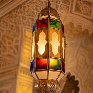 May include: A hanging lantern featuring a metallic frame with intricate cutouts and panels of colored glass in red, blue, green, and yellow, alongside frosted white glass sections. The fixture is suspended by a metal chain from the ceiling.