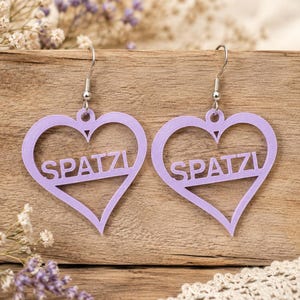 Peut inclure: Une paire de boucles d'oreilles pendantes en forme de cœur violet clair avec le mot SPATZI découpé au centre. Les boucles d'oreilles sont fixées à des crochets en métal argenté et présentent une finition mate et un design ajouré.