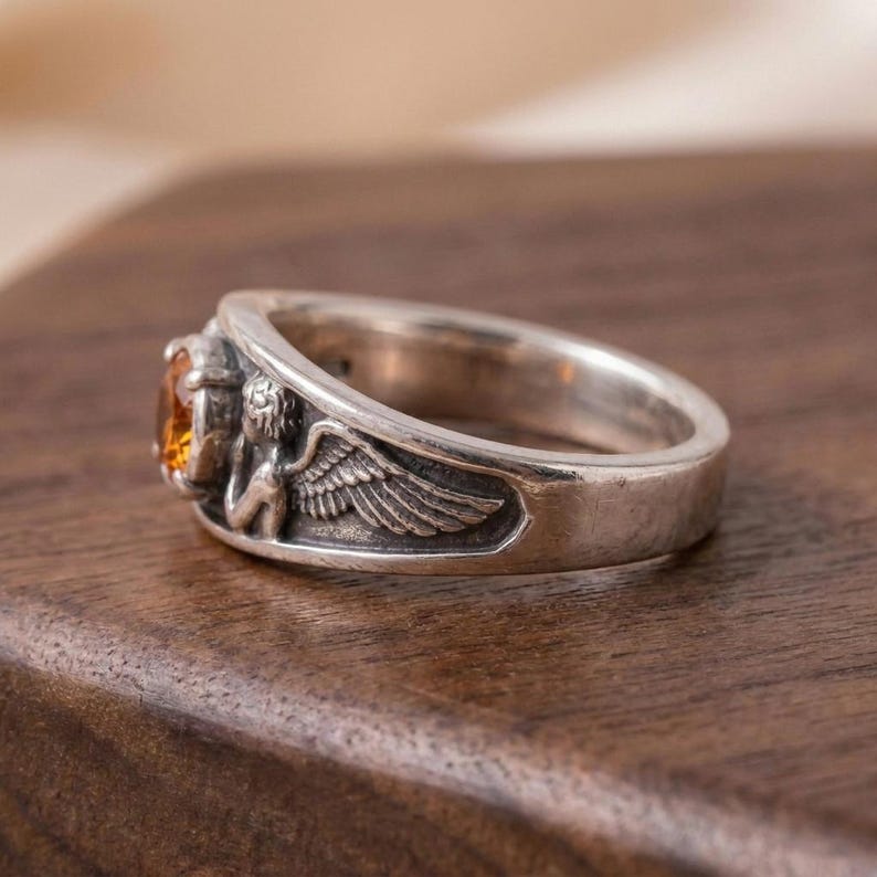 May include: A silver ring with an oval, amber-colored gemstone. The ring features a detailed relief of an angel with wings on one side. The ring is set on a wooden surface.
