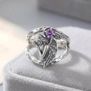 K&ouml;nnte beinhalten: Ein silberner Ring mit einer skulptierten Engelsfigur, deren Fl&uuml;gel sich entlang der Ringschiene erstrecken. Der Engel ist in einer gebeugten Haltung dargestellt, und ein einzelner runder, facettierter violetter Edelstein ist im oberen Teil des Rings eingefasst.