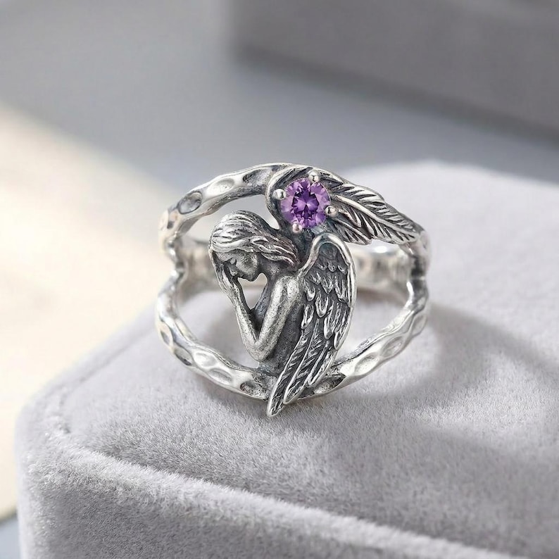 K&ouml;nnte beinhalten: Ein silberner Ring mit einer skulptierten Engelsfigur, deren Fl&uuml;gel sich entlang der Ringschiene erstrecken. Der Engel ist in einer gebeugten Haltung dargestellt, und ein einzelner runder, facettierter violetter Edelstein ist im oberen Teil des Rings eingefasst.