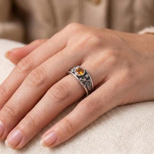 May include: A silver ring with an oval, yellow gemstone. The ring has an ornate design with detailed carvings on the band. The gemstone is set in a bezel setting, and the ring is worn on a finger.