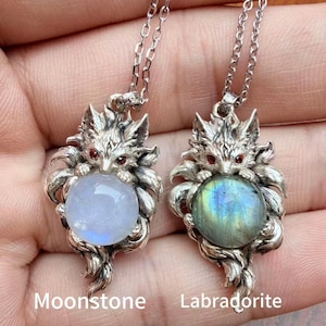 May include: Two silver fox pendants on chains, each featuring a round gemstone held by the fox. The left pendant contains a translucent moonstone, while the right pendant holds a labradorite with blue and green iridescence. Both foxes have small red eyes.