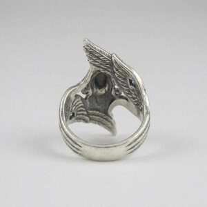 May include: A silver-toned metal ring featuring an intricate design of two wings that curve upward to meet at the top. The surface of the metal has a darkened, oxidized finish in the recessed areas to highlight the detailed feather textures.