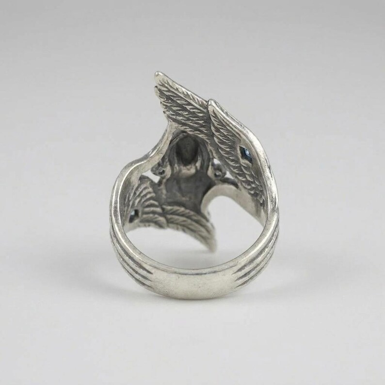 May include: A silver-toned metal ring featuring an intricate design of two wings that curve upward to meet at the top. The surface of the metal has a darkened, oxidized finish in the recessed areas to highlight the detailed feather textures.