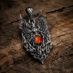 May include: A silver-toned metal pendant featuring a winged figure with outstretched wings positioned above a central oval-shaped orange gemstone. The lower portion of the pendant is sculpted into detailed flame-like patterns, and a bail is at the top.