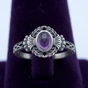 May include: A silver ring with an oval, purple gemstone. The gemstone is surrounded by a decorative floral design. The band features an intricate pattern. A classic piece of jewelry.