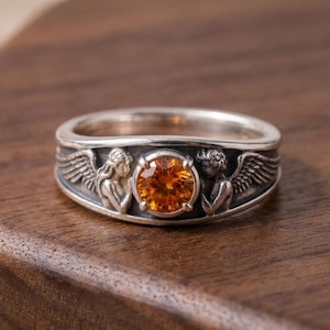May include: A silver ring featuring a large, round, orange gemstone. The band is detailed with two angel figures with wings on either side of the gemstone. The ring is set against a dark wood surface.