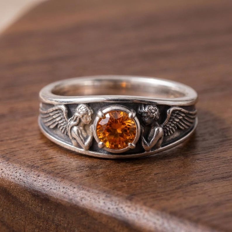 May include: A silver ring featuring a large, round, orange gemstone. The band is detailed with two angel figures with wings on either side of the gemstone. The ring is set against a dark wood surface.