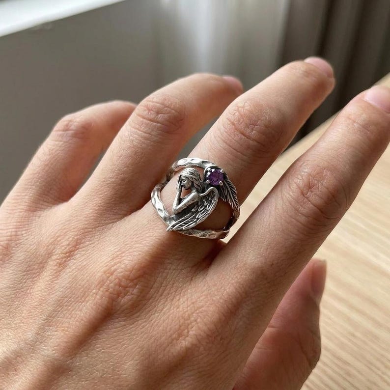 K&ouml;nnte beinhalten: Ein silberner Ring mit einer skulptierten Engelsfigur, deren Fl&uuml;gel sich um die Ringschiene legen. Das Metall hat ein oxidiertes Finish mit einer geh&auml;mmerten Textur. Ein kleiner, runder, violetter Edelstein ist in die Ringschiene nahe dem Fl&uuml;gel des Engels eingefasst.