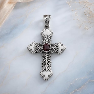 Peut inclure: Un pendentif croix en argent avec un travail de métal complexe, quatre incrustations en émail blanc en forme de trèfle aux extrémités et une pierre précieuse facettée rouge foncé sertie au centre. Il comporte une bélière décorative.