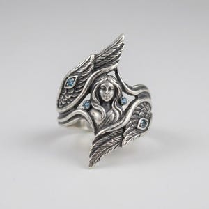 May include: A sterling silver ring featuring a central relief of a female figure with flowing hair, flanked by two detailed wings. The design is accented with four small, round, light blue gemstones embedded within the silver wing structures.