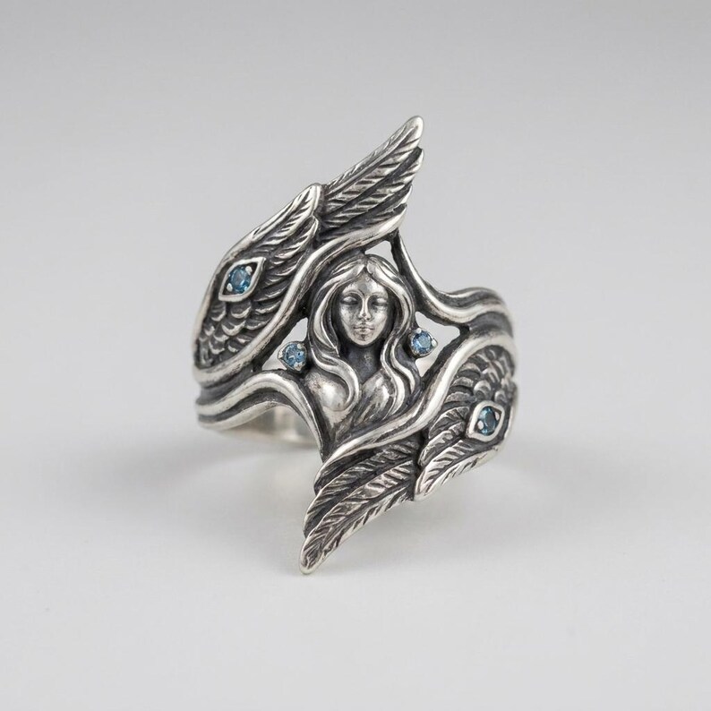 May include: A sterling silver ring featuring a central relief of a female figure with flowing hair, flanked by two detailed wings. The design is accented with four small, round, light blue gemstones embedded within the silver wing structures.