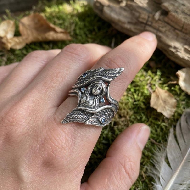 May include: A silver ring featuring a central sculpted figure of a person with long hair surrounded by two large, detailed wings. The design is accented with four small, light blue gemstones embedded within the wings and near the central figure.