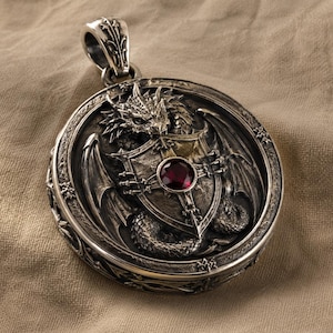 May include: A circular silver pendant featuring a detailed relief of a dragon clutching a shield. The shield is centered with a round, deep red gemstone. The pendant has an ornate bail at the top and intricate engravings along the outer edge.