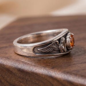 May include: A silver ring with an orange gemstone. The ring features a detailed design of a winged figure. The gemstone is round and has a warm, amber color. The ring is set on a dark wooden surface.