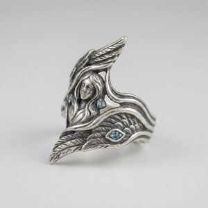 May include: A sterling silver ring featuring a sculpted figure with flowing hair surrounded by detailed wings. The metal has an oxidized finish to highlight the intricate textures, and the design is accented with four small, round, light blue stones.