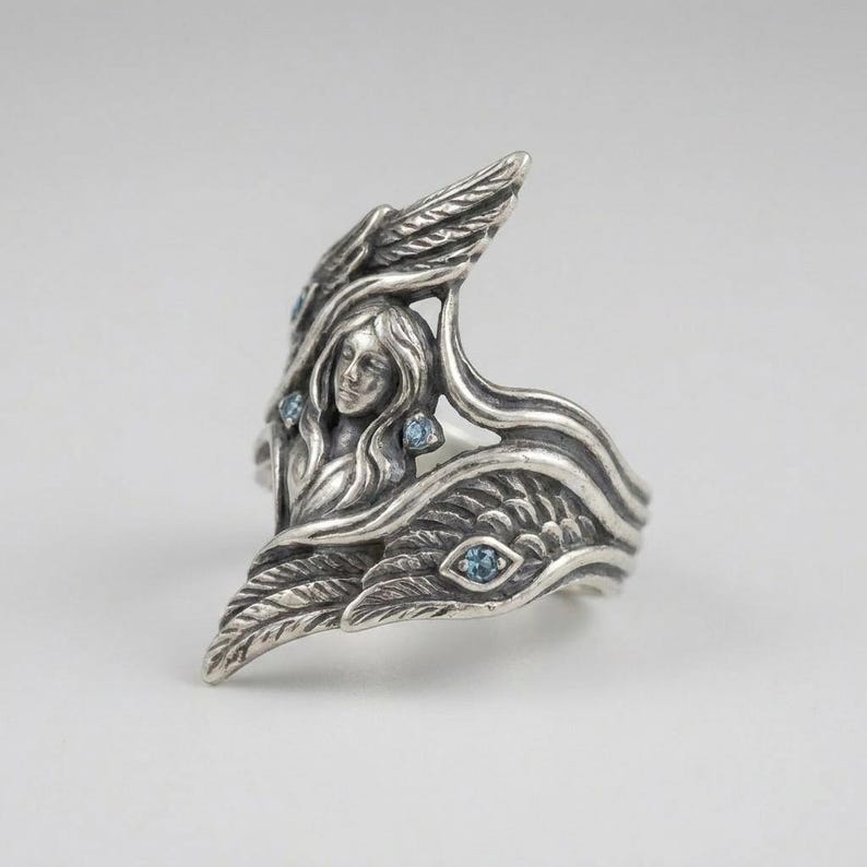May include: A sterling silver ring featuring a sculpted figure with flowing hair surrounded by detailed wings. The metal has an oxidized finish to highlight the intricate textures, and the design is accented with four small, round, light blue stones.