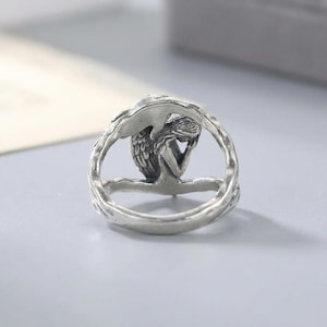 K&ouml;nnte beinhalten: Ein silberfarbener Metallring mit einer skulptierten Figur eines gefl&uuml;gelten Engels mit langem Haar, der in einer sitzenden, nachdenklichen Pose dargestellt ist. Die Ringschiene hat eine strukturierte, organische Optik, die an Zweige erinnert.