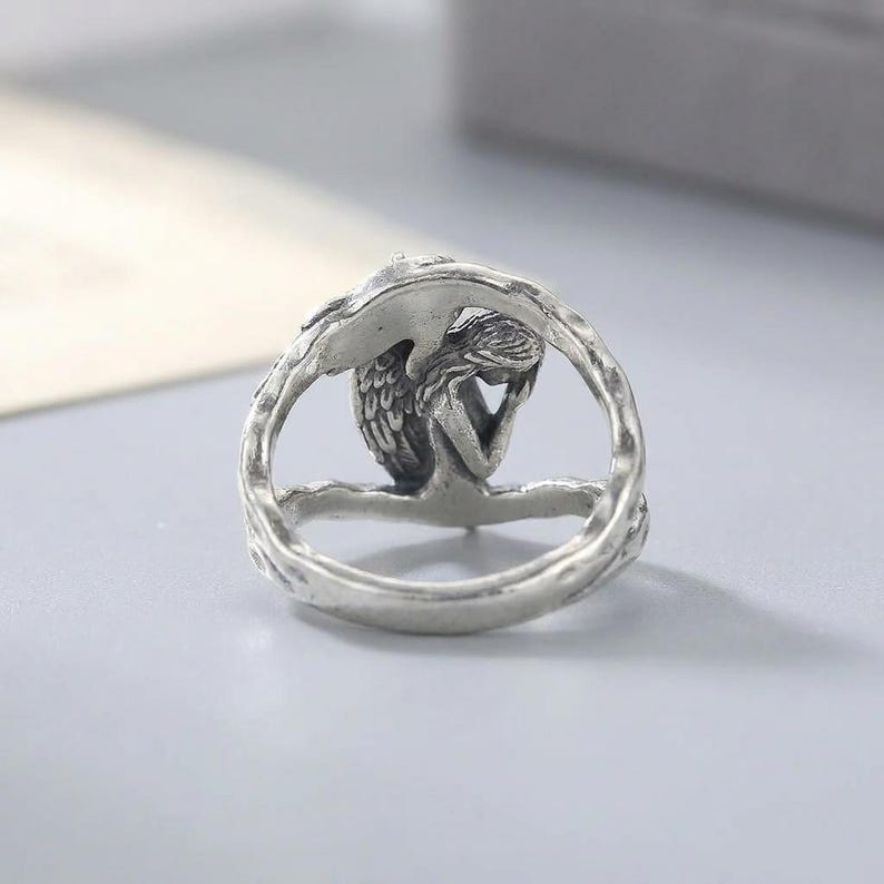 K&ouml;nnte beinhalten: Ein silberfarbener Metallring mit einer skulptierten Figur eines gefl&uuml;gelten Engels mit langem Haar, der in einer sitzenden, nachdenklichen Pose dargestellt ist. Die Ringschiene hat eine strukturierte, organische Optik, die an Zweige erinnert.