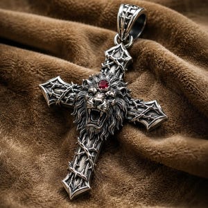 May include: A silver-toned metal cross pendant featuring a detailed lion head at the center with an open mouth and a small red gemstone set on its forehead. The cross is decorated with a raised thorn vine pattern and has a large top bail loop.
