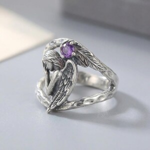 K&ouml;nnte beinhalten: Ein silberfarbener Ring mit einer skulpturalen Engelsfigur mit gefalteten Fl&uuml;geln und einem kleinen, runden, violetten Edelstein an der Oberseite. Die Ringschiene hat eine texturierte, geh&auml;mmerte Optik und ein offenes Design.