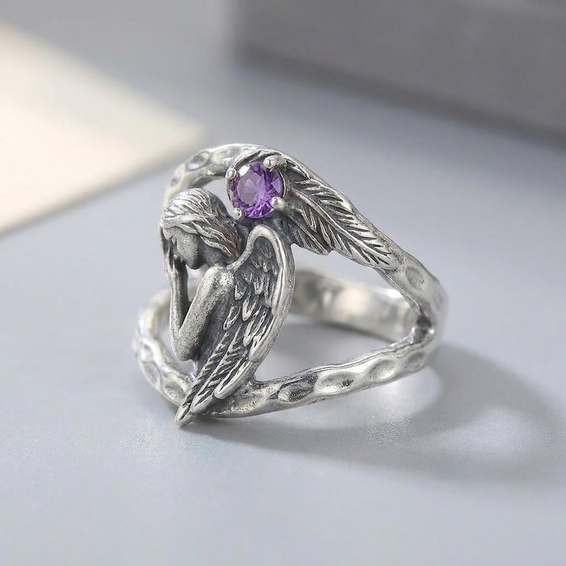 K&ouml;nnte beinhalten: Ein silberfarbener Ring mit einer skulpturalen Engelsfigur mit gefalteten Fl&uuml;geln und einem kleinen, runden, violetten Edelstein an der Oberseite. Die Ringschiene hat eine texturierte, geh&auml;mmerte Optik und ein offenes Design.