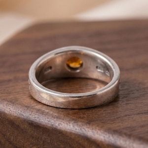 May include: A silver ring with a round, amber-colored gemstone set in the center. The ring has a thick band and is displayed on a dark wooden surface. The gemstone is the focal point, catching the light and adding a touch of color.