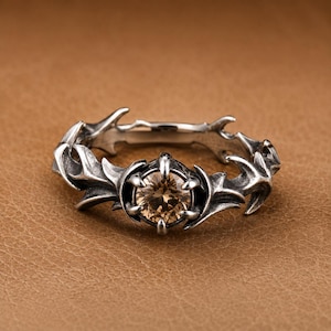 Puede incluir: Un anillo de tono plateado con acabado oxidado y una piedra preciosa redonda de color champán sujeta por cuatro garras. La banda presenta un intrincado trabajo de metal puntiagudo y espinoso que se extiende desde el engaste central.